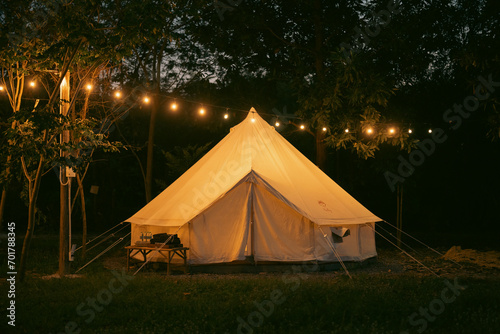 Night and warm light exposure of an illuminated bell tent surrounded by trees and a small camp fire at night, outdoor camping lifestyle