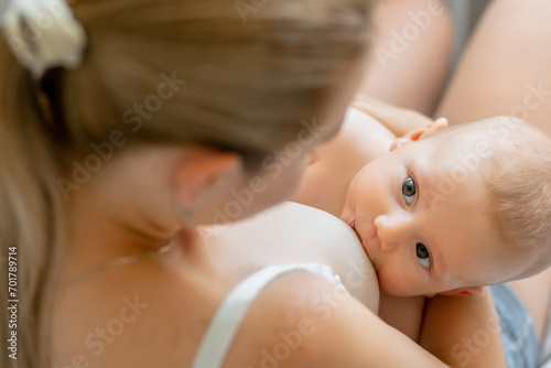 careful young mom sitting on sofa bed and breastfeeding baby while holding son in arms photo