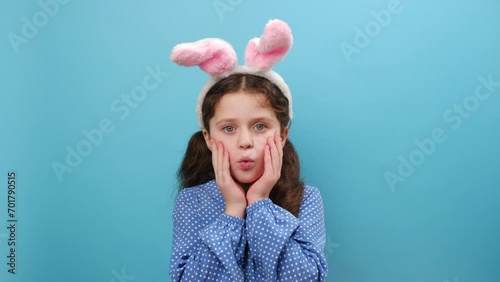 Portrait of preteen girl kid wearing cute easter bunny ears, afraid and shocked looking at camera with surprised expression, fear and excited face, posing isolated over blue color background in studio photo
