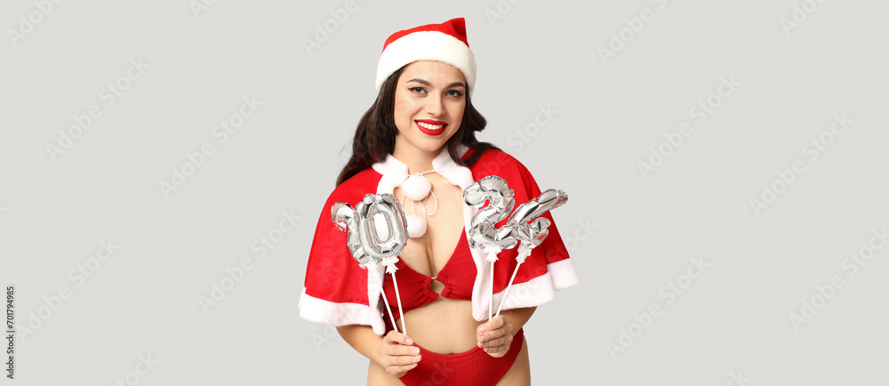 Obraz premium Sexy young woman in lingerie and Santa hat holding balloons in shape of figure 2024 on grey background