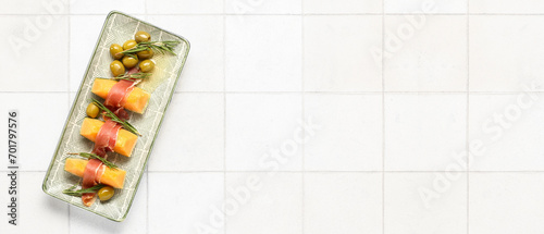 Fototapeta Naklejka Na Ścianę i Meble -  Plate with tasty melon, prosciutto, rosemary and green olives on light tile background with space for text