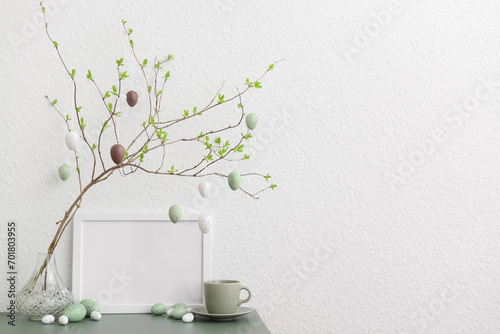 Tree branch with hanging Easter eggs, cup and blank frame on table near white wall photo