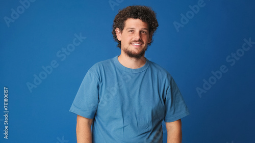 Smiling guy with curly hair dressed in blue t-shirt looking at the camera and waving his hand in welcoming gesture, saying hello to someone, isolated on blue background in the studio