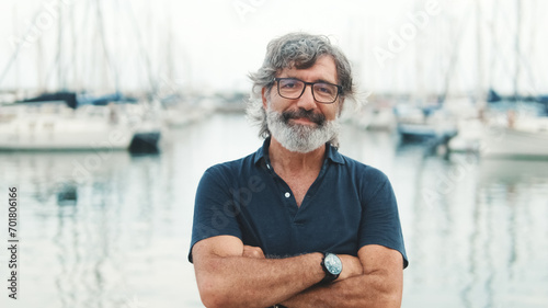 Happy male seigneur in glasses crossing his arms while standing in the port and looking at the camera on yachts background, camera is moving closer photo