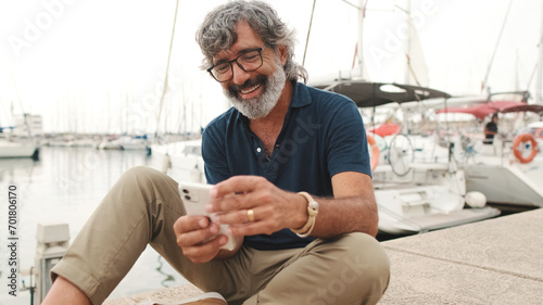Close up, happy senior man in glasses sits in the port on yacht background uses smartphone, looks at funny video, scrolling on the phone screen, communicates in social networks photo