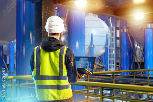 Man industrial engineer. Guy on territory of plant. Industrialist with laptop with back to camera. Oil refinery engineer. Man stands on industrial mezzanine. Industrial engineer near oil tanks photo