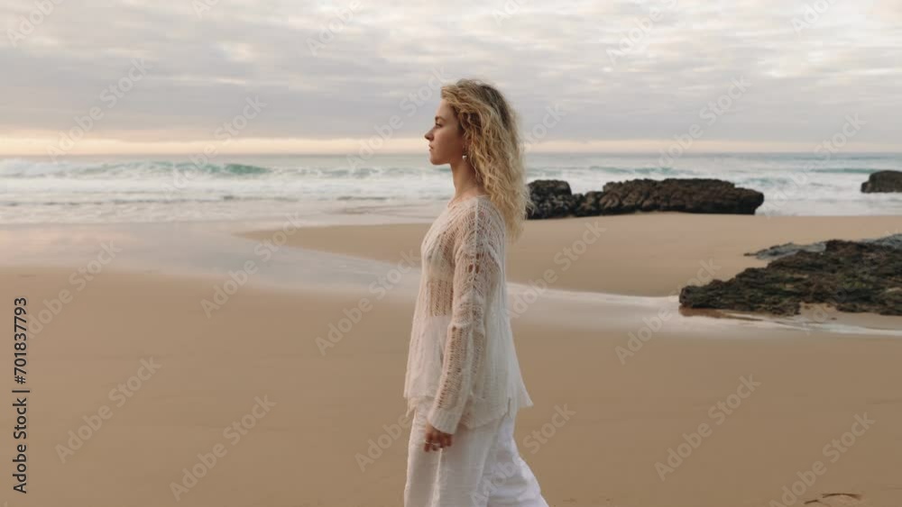 A beautiful young blonde woman in white walks alone on an empty beach, deep in pensive thought during an amazing sunset A beautiful young blonde woman in white walks alone on an empty beach, deep in pensive thought during an amazing sunset