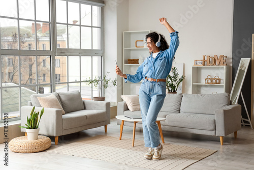 Young African-American woman in headphones with mobile phone dancing at home photo