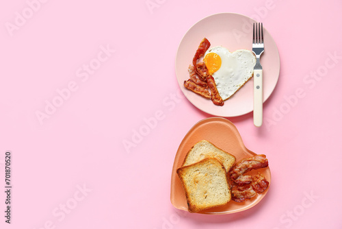 Plates with tasty bacon, heart made of fried egg and toasts on pink background