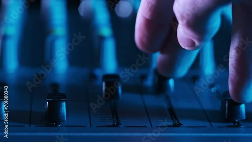 DJ hand adjusts settings volume level controls on mixing console faders in neon light close-up. Sound engineer moves knobs up and down on audio mixer. Man working in colorful background plays music photo