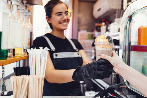 Smiling young barista girl in casual clothes, apron and black gloves gives cocktail with cream and cookies to her customer.