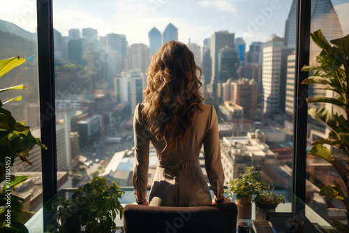 Businesswoman stands by a window in a high-rise office, overlooking a dusk-lit cityscape, her desk adorned with personal mementos. photo