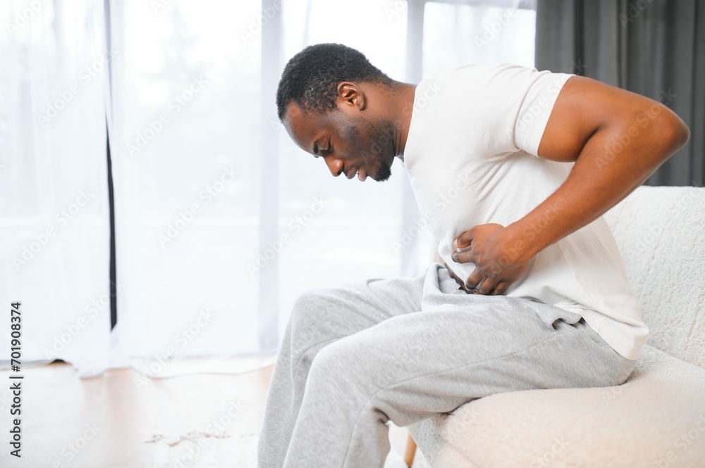 African american guy having stomach ache after eating touching aching stomach suffering from pain sitting on sofa at home.