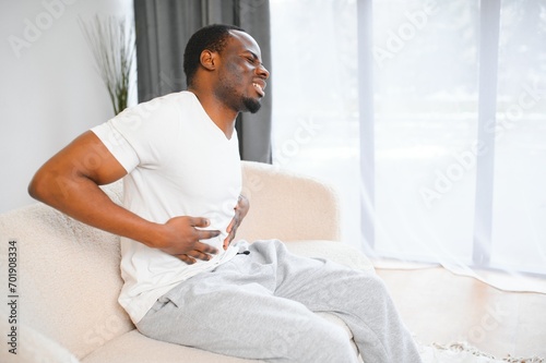 African american guy having stomach ache after eating touching aching stomach suffering from pain sitting on sofa at home.