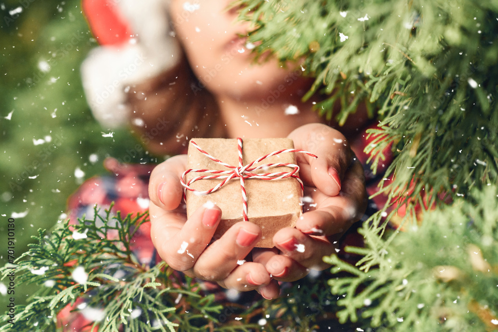 Φωτογραφία Woman in a santa claus red hat holds out small gift through green fir tree branches in the forest during snowfall at winter