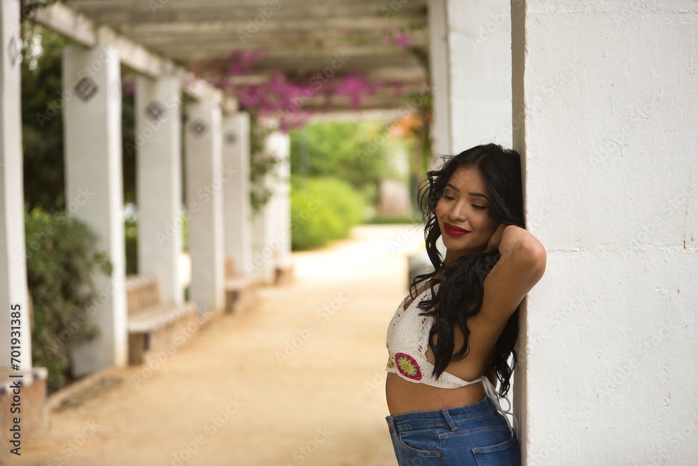 South American woman, young, beautiful, brunette, with crochet top and jeans leaning on a column, with a tender and dreamy look. Concept of beauty, fashion, trend, ethnicity, diversity. South American woman, young, beautiful, brunette, with crochet top and jeans leaning on a column, with a tender and dreamy look. Concept of beauty, fashion, trend, ethnicity, diversity.