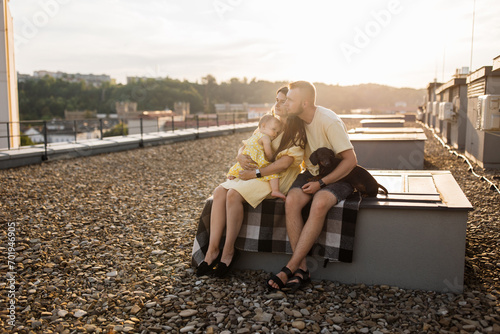 Side view of family of three with dachshund dog sitting on blanket on open space. Surprised mother showing something to little daughter outdoors. Happy spouse enjoying scenery and sunset together.