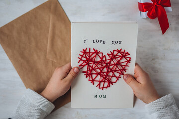 Concept of children craft, handmade gift for Mother's Day. Little boy holding craft card with embroidering in the shape of heart and printed message I love you Mom. Kid's leisure activity, handcraft