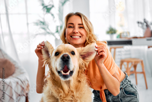 Fun with animal. Carefree woman joyfully lifting ears of adorable golden retriever and smiling at camera while sitting on floor. Young pet owner enjoying every moment spending with her best companion.