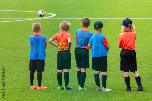Young football players in colourful shirts stand in a row on a green football field in front of the ball. Children's football school