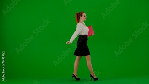 Portrait of attractive office girl on chroma key green screen. Woman in skirt and blouse walking cutely with red paper folder, smiling expression. © kinomaster