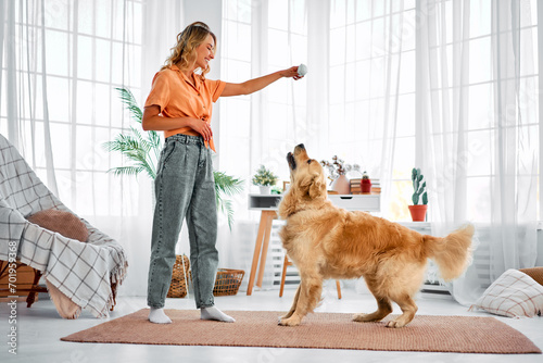 A beautiful stylish woman in jeans and an orange shirt is training her dog at home. A beautiful fluffy dog is running after his favorite toy.