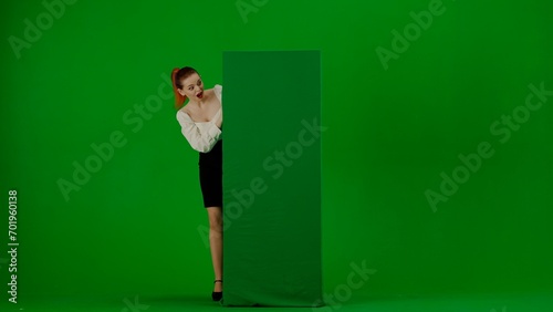 Portrait of attractive office girl on green screen with chromakey. A woman in a skirt and blouse peeks out from behind a advertising billboard, smiling with an amazed face. © kinomaster