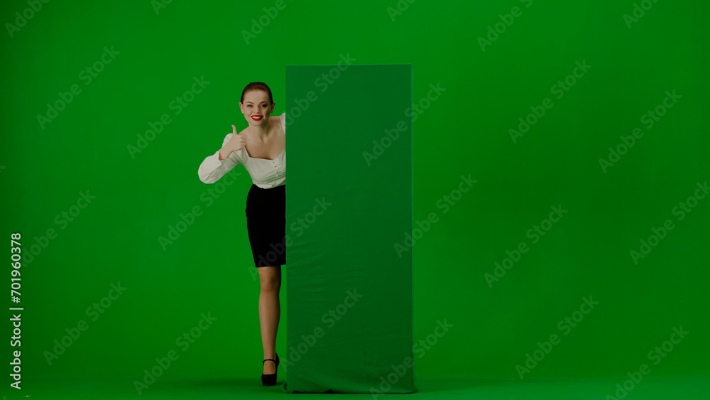 Portrait of attractive office girl on green screen with chromakey. Woman in a skirt and blouse looks out from behind a advertising billboard, smiles and points a thumbs up