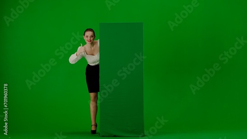 Portrait of attractive office girl on green screen with chromakey. Woman in a skirt and blouse looks out from behind a advertising billboard, smiles and points a thumbs up © kinomaster