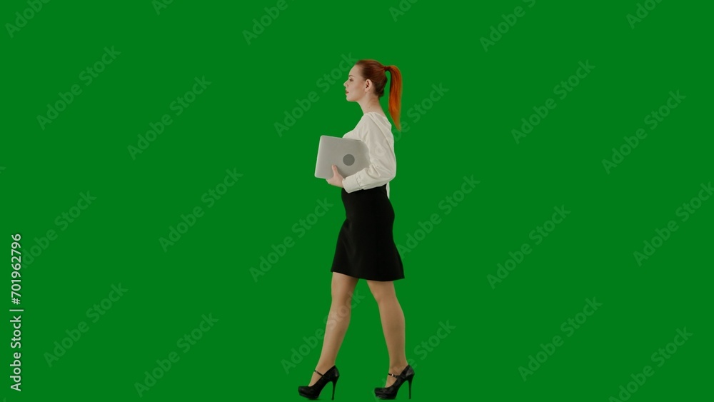 Portrait of attractive office girl on chroma key green screen. Woman in skirt and blouse walking holding laptop. Side view.