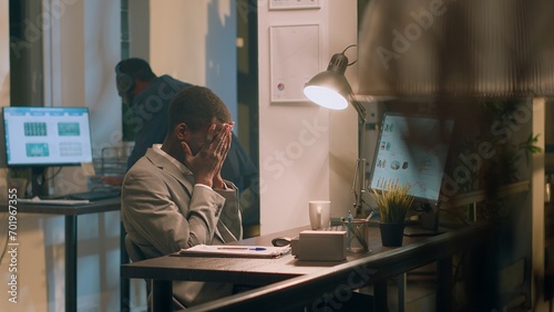 African american accountant exasperated with noisy colleague listening to music and pretending to beat drums during nightshift. Employee annoyed by distracting coworker banging desk in office photo