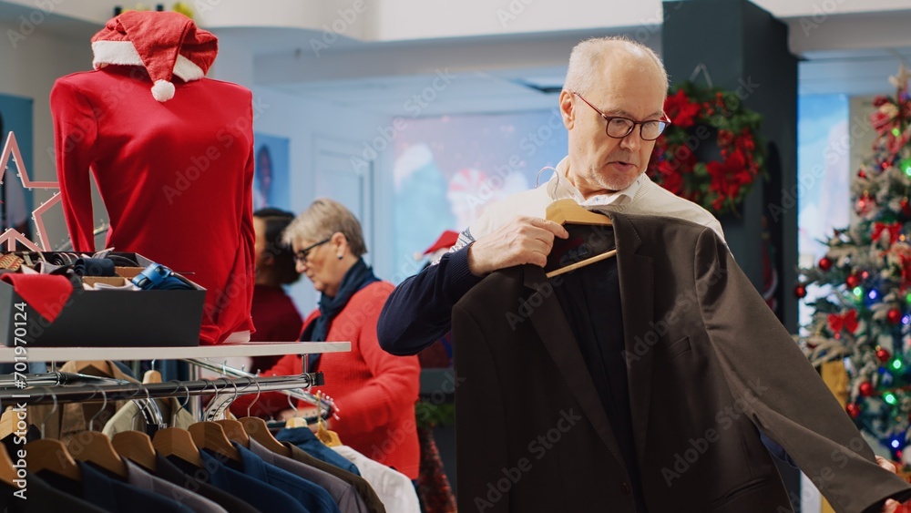 Senior man browsing blazers in festive clothing store during winter holiday season, looking at formal attire pieces, doing Christmas shopping spree in xmas decorated shop