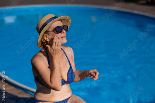 Portrait of an old woman in a straw hat, sunglasses and a swimsuit applying sunscreen to her face while relaxing by the pool. 