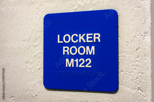 Blue LOCKER ROOM M122 sign with braille, on a white block wall. 