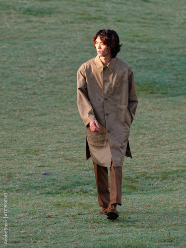 Portrait of a handsome Chinese young man in khaki overcoat with hand in pocket walking on green grass field in sunny day, male fashion, cool Asian young man lifestyle.
