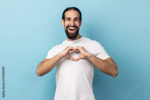 I love you sign. Be my valentine. Portrait of romantic man with beard wearing white T-shirt shapes heart confesses in love, looking at camera. Indoor studio shot isolated on blue background. photo