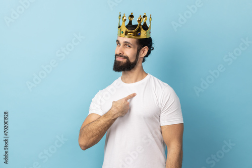 Portrait of proud man with beard wearing white T-shirt and golden crown and pointing himself, looking at camera with smile, superior privileged status. Indoor studio shot isolated on blue background.