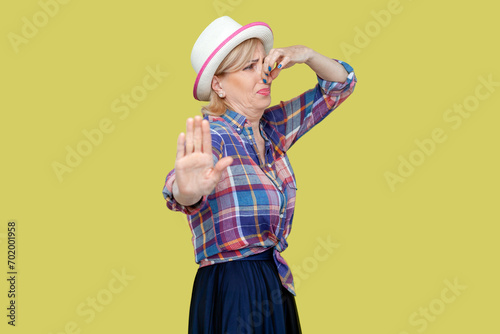 Senior woman wearing checkered shirt and hat covers nose with hand, smells something awful, pinches nose, frowns in displeasure, makes stop gesture. Indoor studio shot isolated on yellow background. photo