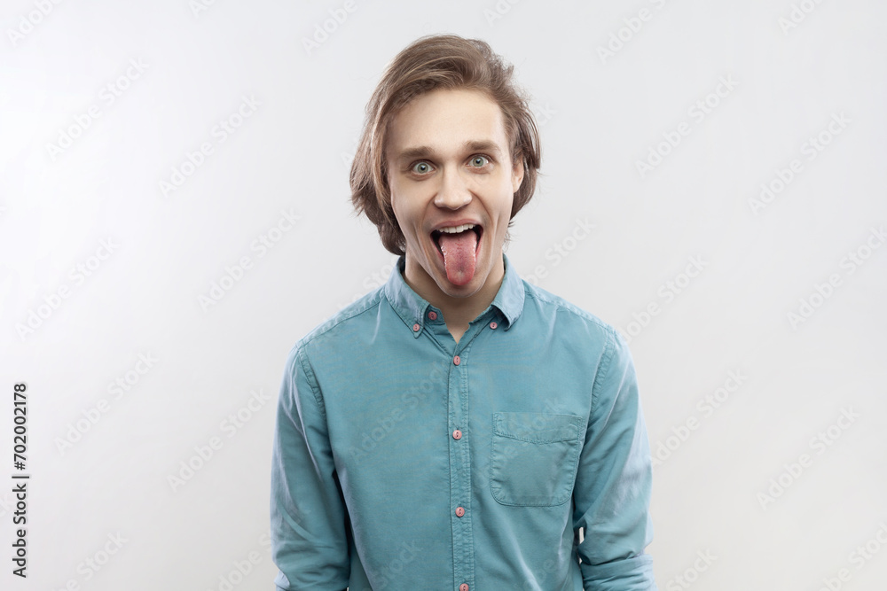 Portrait of funny crazy childish attractive young man standing looking at camera and sticking tongue out, having fun, wearing blue shirt. Indoor studio shot isolated on gray background. Portrait of funny crazy childish attractive young man standing looking at camera and sticking tongue out, having fun, wearing blue shirt. Indoor studio shot isolated on gray background.