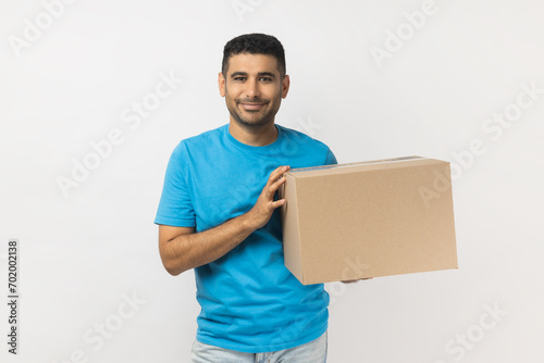 Portrait of smiling cheerful unshaven man wearing blue T- shirt standing holding cardboard box with his stuff, relocating to a new apartment. Indoor studio shot isolated on gray background.
