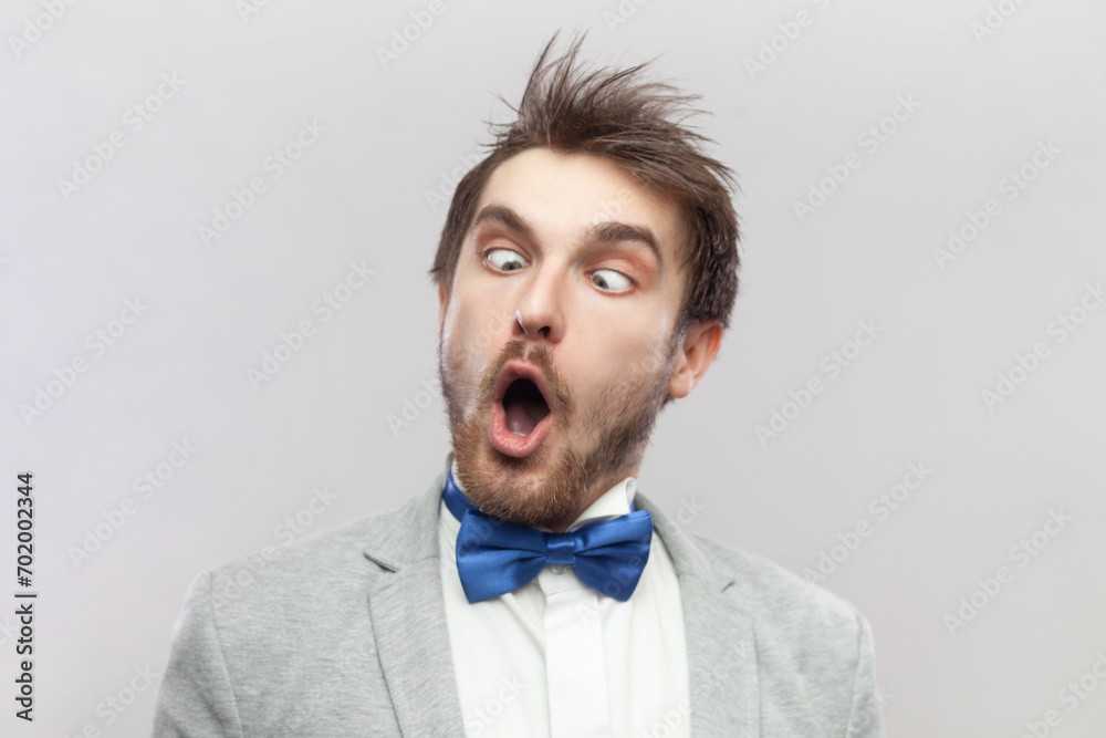 Portrait of funny crazy childish bearded man keeps mouth open and eyes crossed, making foolish face, wearing grey suit and blue bow tie. Indoor studio shot isolated on gray background. Portrait of funny crazy childish bearded man keeps mouth open and eyes crossed, making foolish face, wearing grey suit and blue bow tie. Indoor studio shot isolated on gray background.