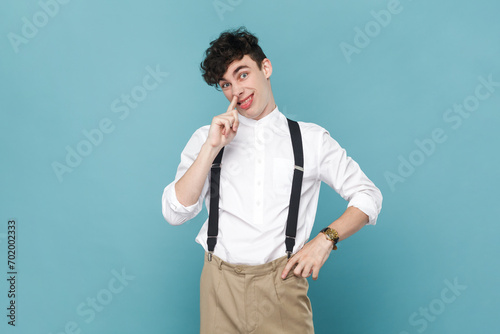 Portrait of mischievous man in shirt and suspender putting finger into his nose and showing tongue, fooling around, bad habits, disrespectful behavior. Indoor studio shot isolated on blue background.