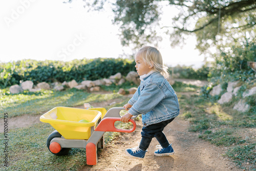 Little girl with a toy wheelbarrow walks along a dirt path in the park photo