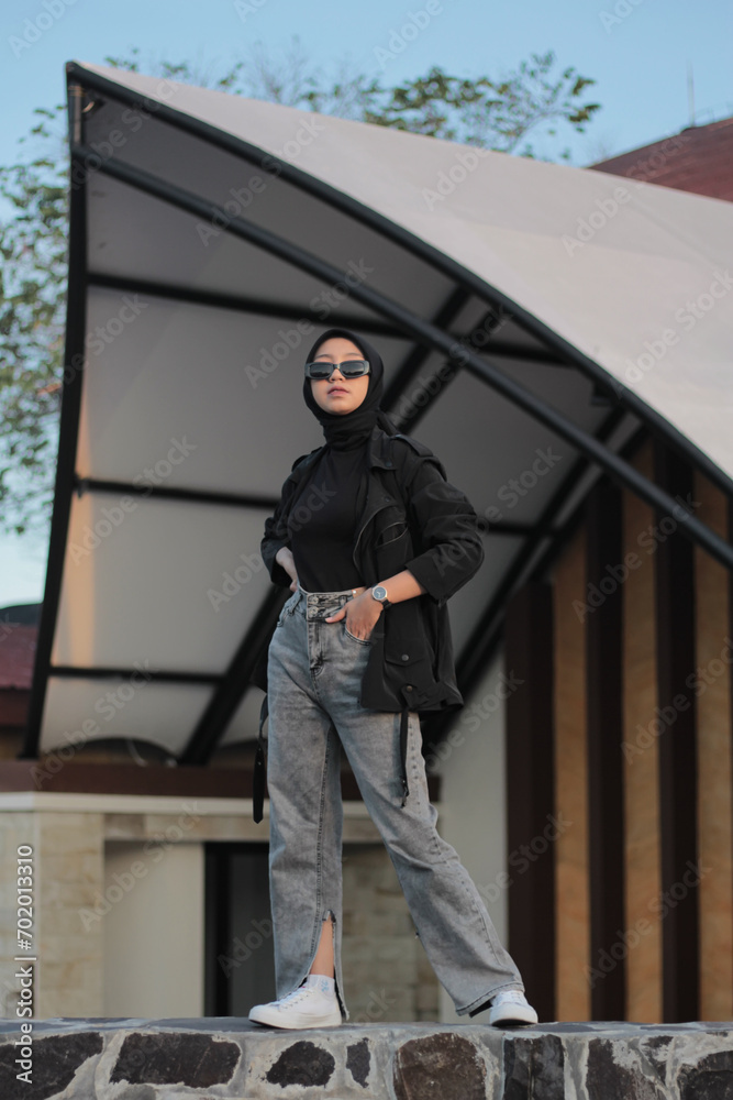 A young Asian woman wearing a hijab and jeans posing outdoors, following a concept with a black outfit