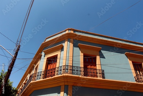 
A vibrant blue building with orange trim and ornate balconies under a clear blue sky, crisscrossed by power lines.