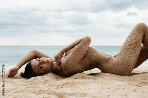 Gorgeous young woman lying in the sand, her body artfully covered with grains of sand, on a beach photo