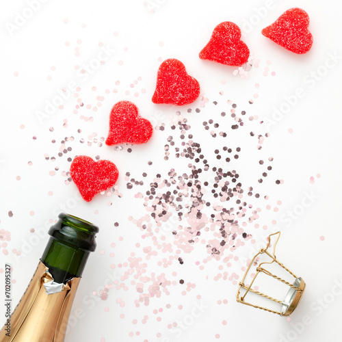 Closeup flatlay of a sparkling wine bottle and red candy hearts on a white backgrounf with pink glittering confetti. Valentine's Day decoration in square