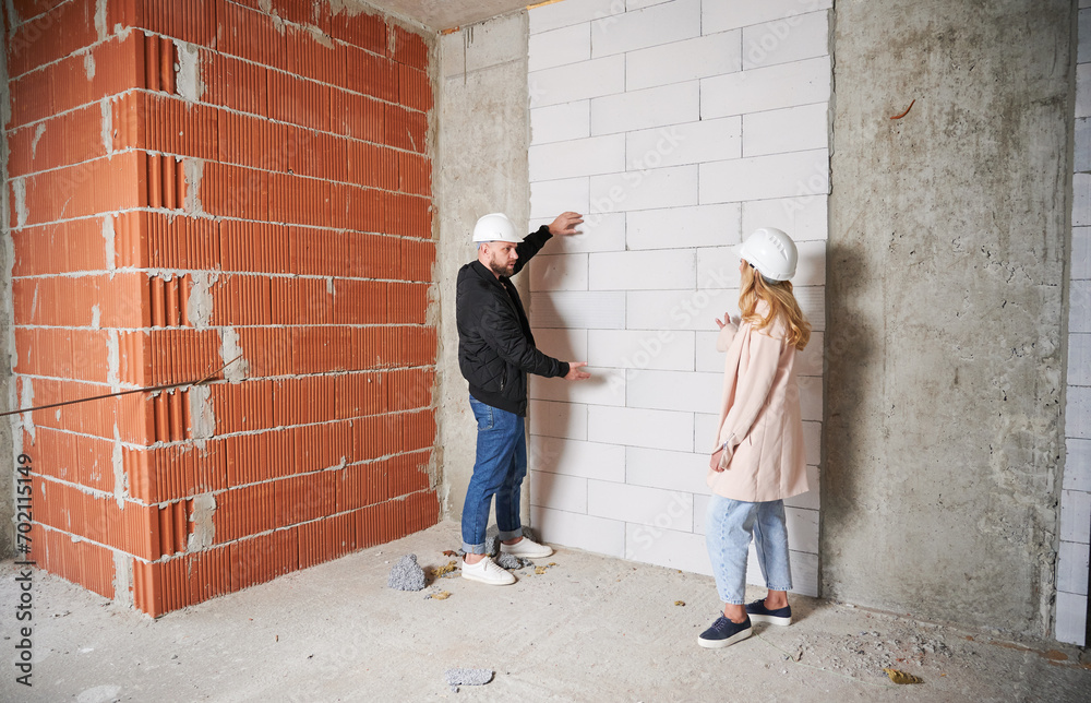 Full length of woman standing by brick wall and discussing apartment replanning and renovation with specialist. Female homeowner having meeting with builder in building under construction.