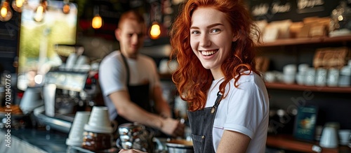 Beautiful Caucasian rad haired lady looks smiling at camera offers hot coffee at modern coffee shop barista posing near counter wearing white t shirt and apron. with copy space image © vxnaghiyev
