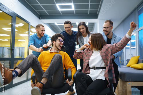 A group of young colleagues are having fun in the office corridor. Young business people ride on office chairs and laugh. Concept of teamwork, relaxation or fun.